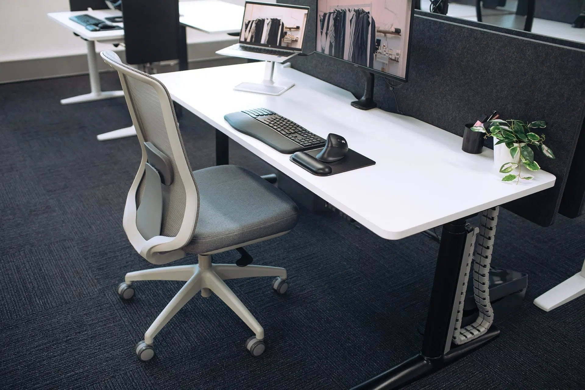 A modern white adjustable standing desk with a black metal frame, paired with a gray mesh office chair, in an office setting.