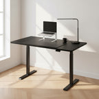 Cloud Office Studio 140cm black dual-motor standing desk with matte finish, laptop stand, and lamp in a bright office.